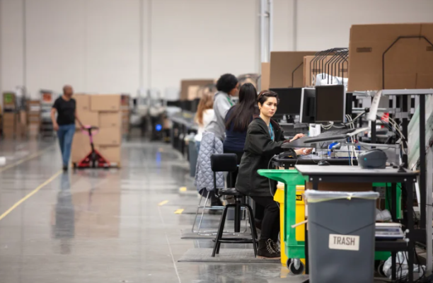 1 The inside of a 3PL shipping bay. A man moving a pallet of boxes with a hand jack is in the background while a woman at a computer coordinating business is in the foreground.