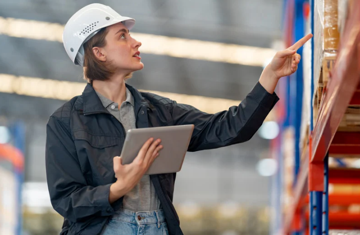 FOX-Doc-07-16-2025_05_35_PM A woman wearing a white hard hat in a 3PL warehouse looking at items with a tablet in her hand.
